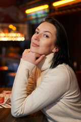 beautiful smiling girl with shoulder-length dark hair sits in a cafe, propping her head with both hands