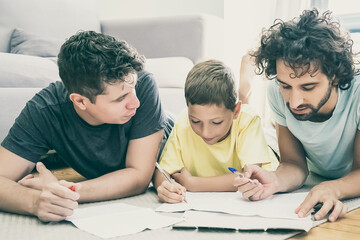 Couple of homosexual parents helping focused boy with school home task, lying on floor at home, writing or drawing in papers. Family and gay parents concept