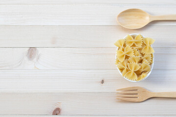 Farfalle in a white cup with spoon and fork on light pine wood background.