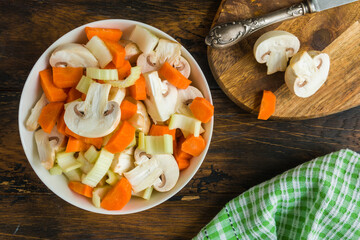 Sliced vegetables, carrot, celery and mushrooms. White bowl, knife and cutboard on wooden rustic table, top view - 408802742