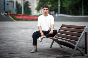 White casual t-shirt mockup on a guy sitting against a blurred park background, on a bench, front...