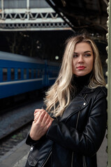Portrait of a posing young showy blonde with long hair in a leather jacket with a red handbag at the train station