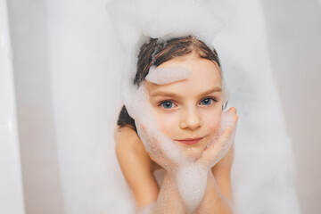 A little girl with long hair, the child is washed, sitting in a white bath with foam from soap, shampoo. Model photography. Face close-up.