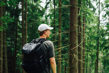 Young male hiker stands with his back in the woods with a backpack on his back and looks away with a serious face. tourist is resting on a hike.