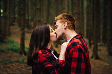 Portrait of a beautiful young man and woman kissing in the woods on a background of coniferous tree trunks. Love story photo, a couple in red shirts hugging and kissing on a hike.