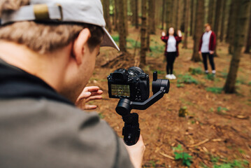 Video maker with a camera on a stabilizer in his hands walks through the woods with a couple of young people and shoots video. Cameraman shoots a couple in love in the untouched forest.