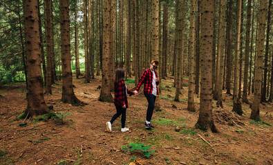 Young couple in red shirt walking through the woods holding hands and smiling faces on. A pair of young hipsters in a coniferous forest holding hands.