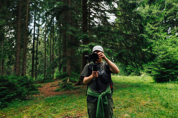 A male videographer with a camera on a stabilizer stands in the woods against a background of trees and adjusts the technique for shooting. Creating video content in nature