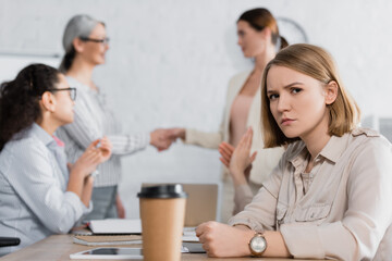 serious businesswoman looking at camera near interracial coworkers on blurred background