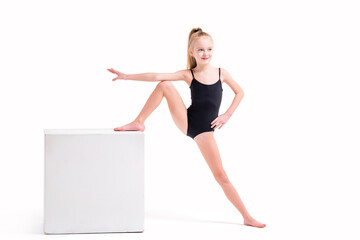 Little girl gymnast in a black swimsuit standing with one foot on white cube, isolated on white background