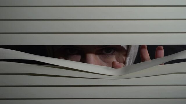 Close up front portrait from indoors of brunette woman in headscarf pushing blinds at window with hands at night. Looking fearfully outside
