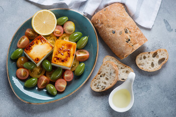 Turquoise plate with baked feta cheese, tomatoes, green olives, ciabatta and olive oil, above view on a light-blue stone background
