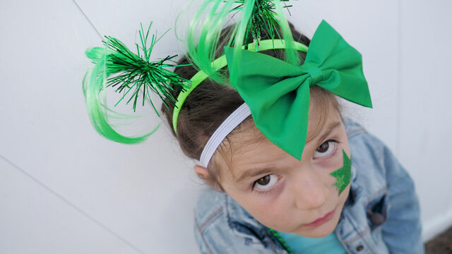 View from above. Cute upset brown hair child with green funny horns, bow and star looking at the camera white wall background, celebrating saint patrick's day.