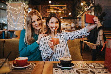 Two best friends sitting in coffee bar or restaurant after shopping and happily talking together. They are using smart phone to take selfie photo.