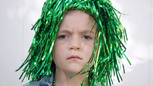Sad girl with brown eyes and frowning eyebrows in green wig looking at the camera celebrating saint patrick's day, white wall background, extreme close up