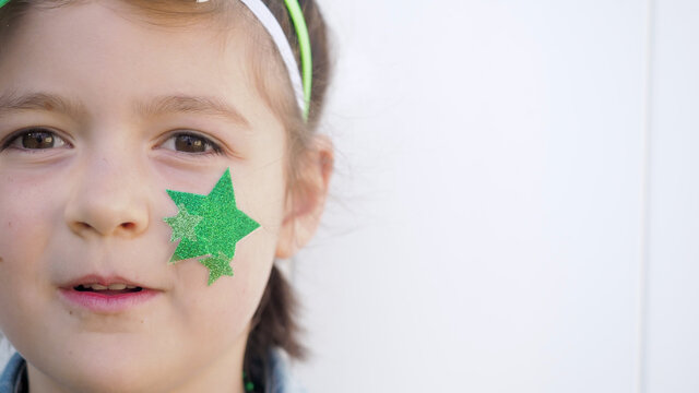 Close up. Cute happy brown hair child with green bow and star on cheek smiling and looking at the camera white wall background, celebrating saint patrick's day.