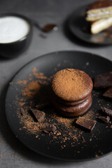 Round chocolate brownie on a black plate and cocoa powder on a gray concrete table. Sweets and coffee background.