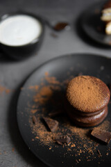 Round chocolate brownie on a black plate and cocoa powder on a gray concrete table. Sweets and coffee background.