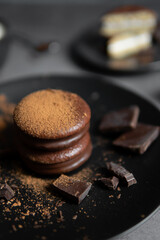 Round chocolate brownie on a black plate and cocoa powder on a gray concrete table. Sweets and coffee background.