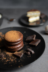 Round chocolate brownie on a black plate and cocoa powder on a gray concrete table. Sweets and coffee background.