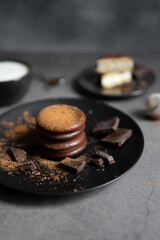 Round chocolate brownie on a black plate and cocoa powder on a gray concrete table. Sweets and coffee background.