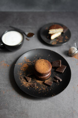 Round chocolate brownie on a black plate and cocoa powder on a gray concrete table. Sweets and coffee background.