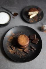 Round chocolate brownie on a black plate and cocoa powder on a gray concrete table. Sweets and coffee background.