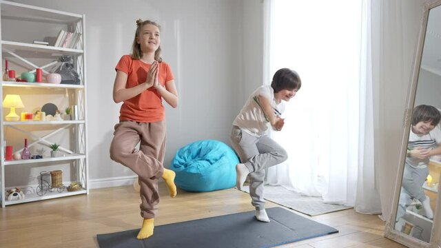 Wide shot portrait of cheerful Caucasian boy and girl standing in tree pose on exercise mat. Joyful brother and sister or friends doing yoga exercise at home. Workout of little yogi.