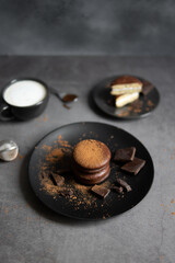 Round chocolate brownie on a black plate and cocoa powder on a gray concrete table. Sweets and coffee background.