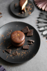 Round chocolate brownie on a black plate and cocoa powder on a gray concrete table. Sweets and coffee background.
