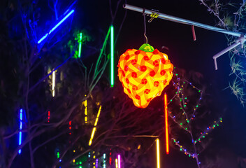 Strawberry shaped lamp hanging with blurred bokeh of colorful lights on the tree at night. A lamp with warm lighting white looks like a heart and strawberry. Concept of love
