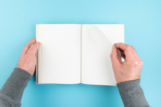 Directly Above View Of Person Turning Page Of Blank White Page Hardcover Book On Blue Background