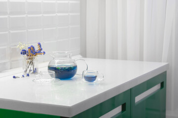 A set of clear glassware teapot and two cups of blue Thai tea in the kitchen interior. Detox tea concept.