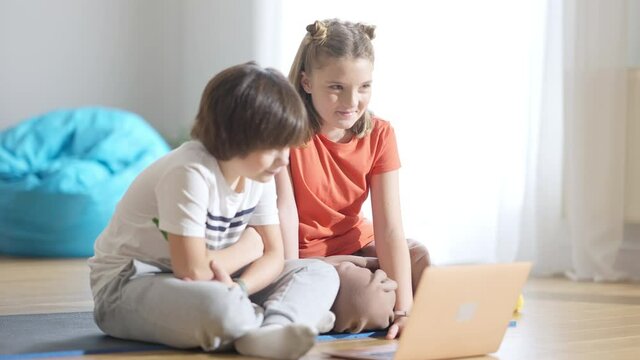 Positive relaxed brother and sister sitting at home using laptop. Portrait of carefree Caucasian boy and girl enjoying leisure indoors. Friends watching movie or cartoons online.