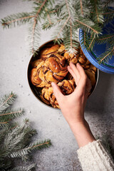 Freshly made Swedish swirl buns with cinnamon, sprinkled with pearled sugar, in tin cookie box with lid open. Surrounded by fir tree branches. Young woman’s hand taking one bun from the box