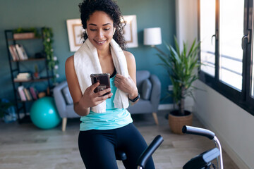 Fototapeta premium Afro young fitness girl using mobile phone while training on exercise bike at home.