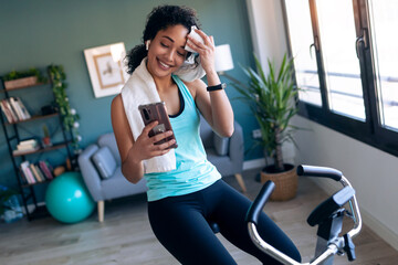 Afro young fitness girl using mobile phone while training on exercise bike at home.