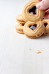 View of woman's hand holding a heart-shaped cookie with strawberry jam, with more cookies on white table with crumbs, in portrait, with copy space