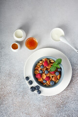 A bowl of freshly made pancake cereal with raspberries, blueberries and fresh mint, shot from above. Jam, honey, sour cream and milk on the table