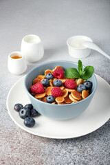 A bowl of freshly made pancake cereal served with fresh raspberries, blueberries and fresh mint. Honey, sour cream and milk on the table behind the bowl