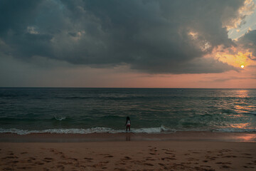 Kobieta na plaży na tle oceanu, zachodzącego słońca i pochmurnego nieba.