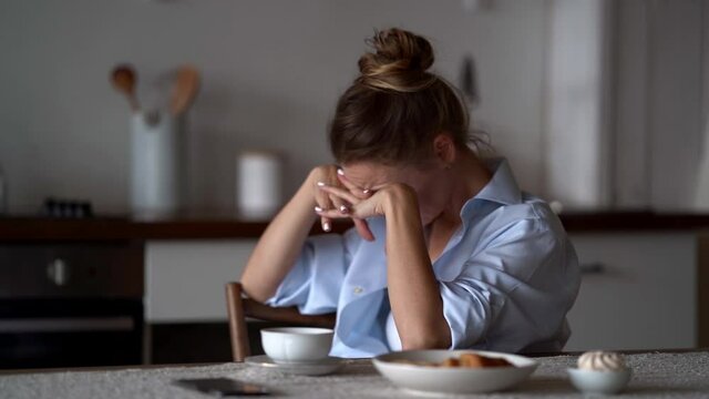 a blonde woman in a white top and blue shirt is sitting at the kitchen table. she puts face down on her clasped hands, then raises head. side view. the average plan