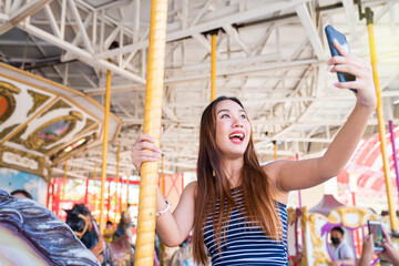 Obraz na płótnie Canvas Young pretty woman taking selfie on carousel in amusement park. Attractive women taking selfie in amusement park.