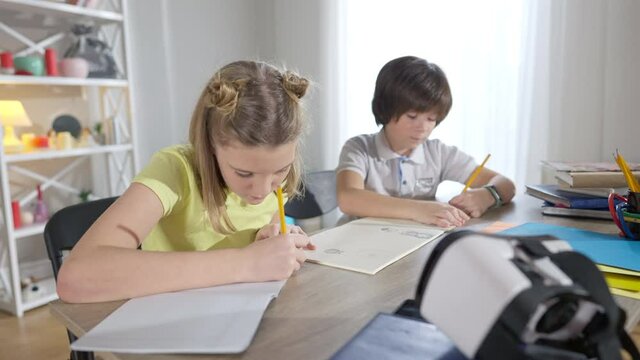 Portrait of focused genius girl thinking and writing in workbook with blurred boy drawing at background. Concentrated intelligent Caucasian children studying in modern classroom at desk. Education.