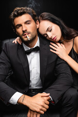 seductive brunette woman leaning on shoulder of man smoking and looking at camera isolated on black