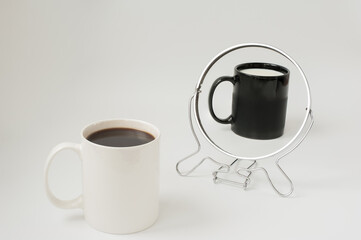A white cup with coffee in front of a mirror are reflected as a black cup filled with milk