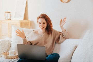 European girl with red hair in a beige sweater sitting on the couch with a laptop
