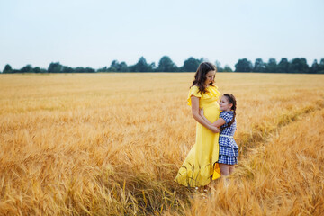 Happy family: a young beautiful pregnant woman with her little cute daughter walking in the wheat orange field on a sunny summer day. Parents and kids relationship. Nature in the country.