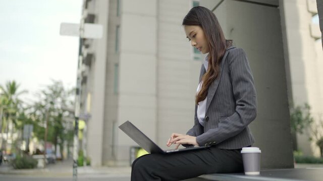Video Slow-motion. A beautiful white-skinned Asian woman in a black suit sitting in front of the office building. Using the laptop on the side of the road in the city.
