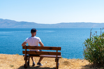 Guy sitting on wooden bench and looking at the blue ocean. Balos close to Chania. Grece